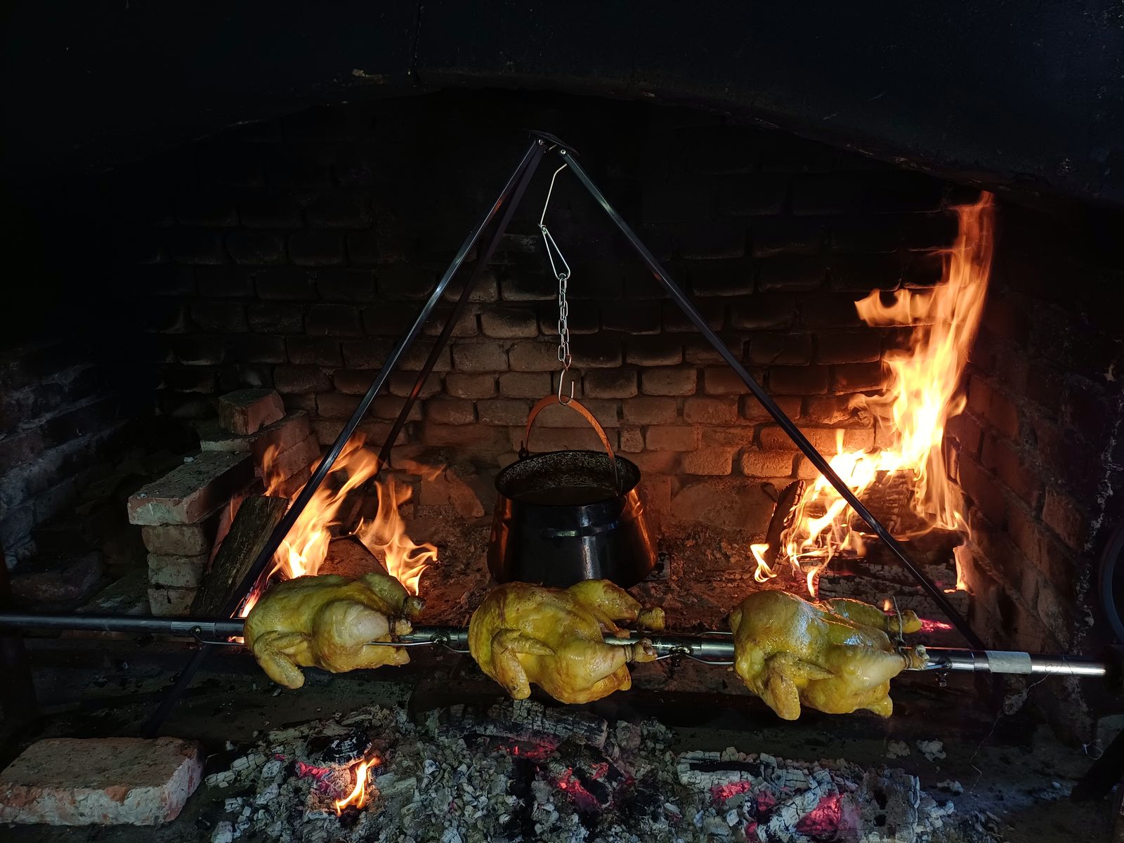 Drei Grillhähnchen am Drehspieß über offenem Feuer, Kessel dahinter